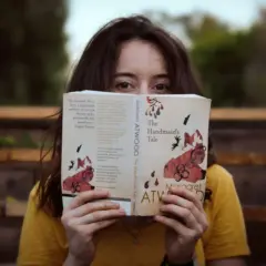 Dziewczyna w żółtej koszulce trzyma przed twarzą otwartą książkę "Opowieść podręcznej" Margaret Atwood.