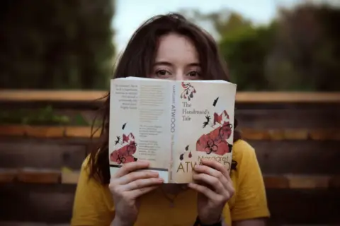 Dziewczyna w żółtej koszulce trzyma przed twarzą otwartą książkę "Opowieść podręcznej" Margaret Atwood.