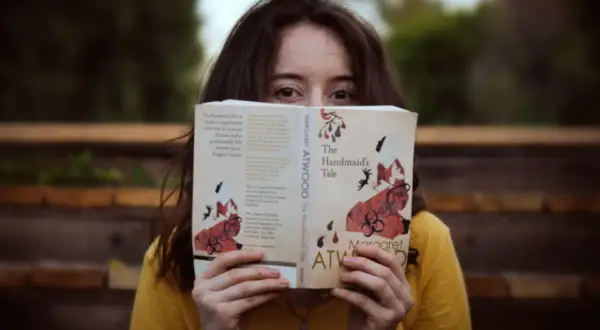 Dziewczyna w żółtej koszulce trzyma przed twarzą otwartą książkę "Opowieść podręcznej" Margaret Atwood.
