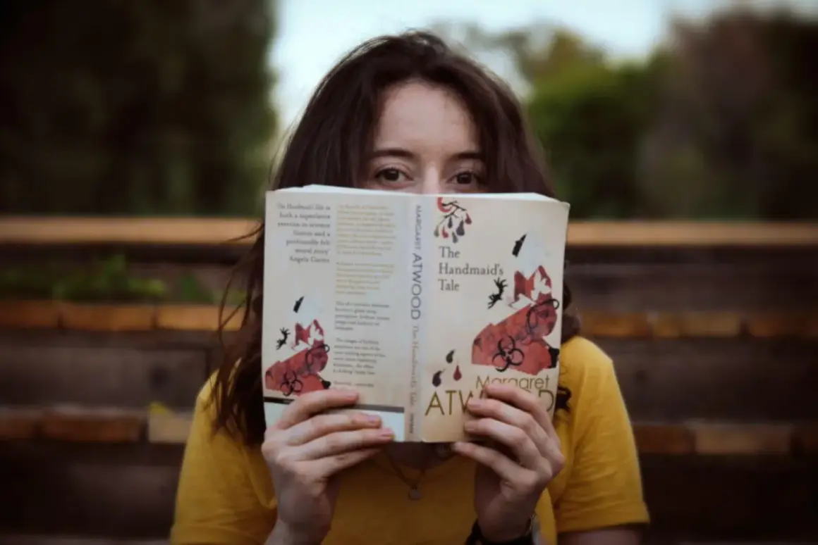 Dziewczyna w żółtej koszulce trzyma przed twarzą otwartą książkę "Opowieść podręcznej" Margaret Atwood.