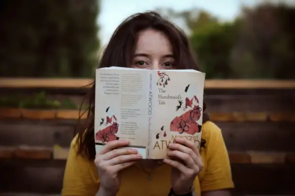 Dziewczyna w żółtej koszulce trzyma przed twarzą otwartą książkę "Opowieść podręcznej" Margaret Atwood.
